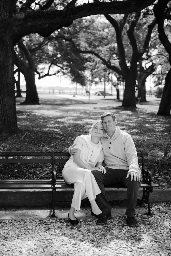 black and white portrait of couple for engagement shoot at the battery in downtown charleston sitting on park bench under trees