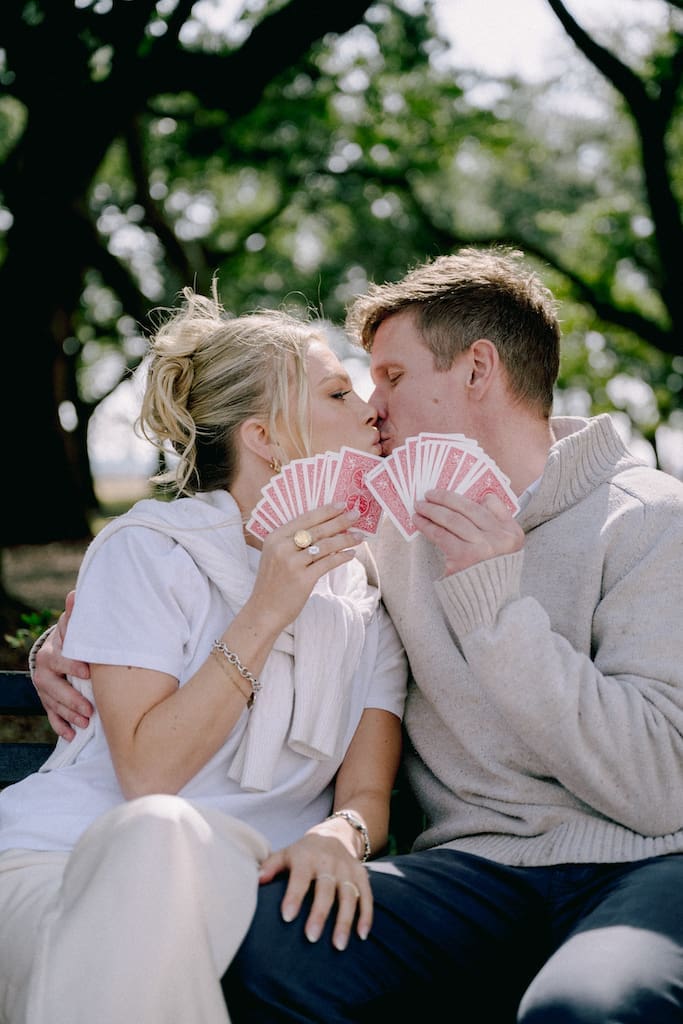 portrait of couple for engagement shoot at the battery in downtown charleston sitting on park bench under trees kissing with cards